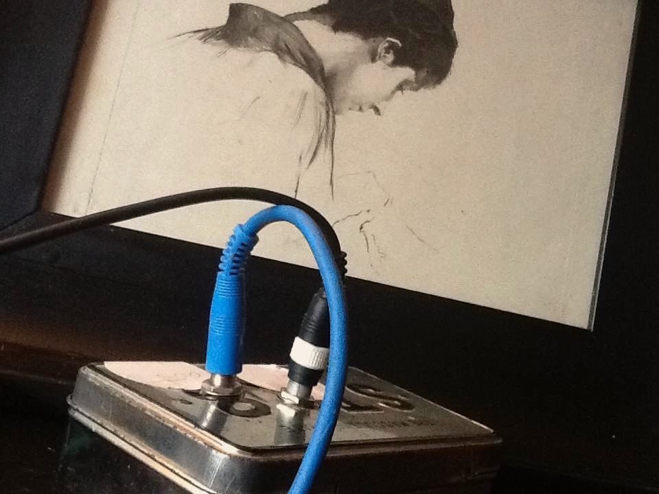One of my old self-built effect boxes: a tin case with two jacks, a blue patch cable and a black cable plugged in, a pencil drawing behind it.